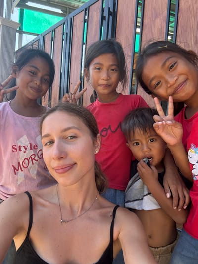 A group of five people, including one adult woman and four children, pose for a selfie outdoors. The woman, with light skin and brown hair pulled back, is in the foreground. She wears a black tank top and a delicate necklace. Behind her, three young girls and one young boy are visible. The girls, with dark hair and eyes, wear t-shirts in pink and red. The boy, also with dark hair and eyes, wears a grey and white shirt and appears to be holding something to his mouth. The background consists of a wooden fence and a glimpse of a green roof structure. The setting is likely Kertasari, Indonesia, given the context provided, suggesting a casual, outdoor gathering. The overall mood is cheerful and friendly, with several individuals flashing peace signs.