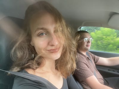 In Sepych, Russia, a close-up selfie captures two individuals inside a vehicle. The woman in the foreground, with light brown hair partially obscuring her face, looks directly at the camera with a slight smile. She is wearing a dark grey top and a delicate gold necklace. A silver earring with three clear beads dangles from her left ear. The man, seated in the passenger seat behind her and slightly out of focus, wears glasses and a brown, ribbed short-sleeved shirt. He appears to be looking ahead, possibly driving. The interior of the car, including the dashboard and seatbelts, is visible. Outside the car window, lush green trees blur due to motion, indicating the vehicle is in transit. The lighting suggests an overcast day.