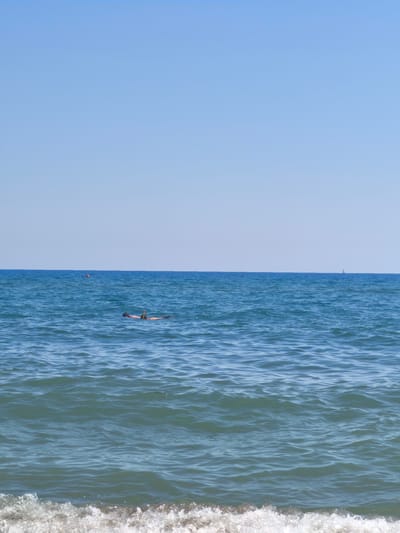 The image depicts a tranquil scene of the sea at Castiglione della Pescaia, Italy, under a clear blue sky. In the middle ground, a person is seen floating on their back, wearing a snorkel mask and fins, suggesting they are engaged in snorkeling or leisurely floating. The water is a deep blue, with gentle waves rippling across its surface. A small orange buoy is visible in the distance to the left, and a distant sailboat can be spotted on the horizon to the right. The foreground features the edge of a breaking wave with white foam washing onto what appears to be a sandy shore, though the shore itself is not clearly visible in this frame. The overall impression is one of a calm and sunny day at the beach.