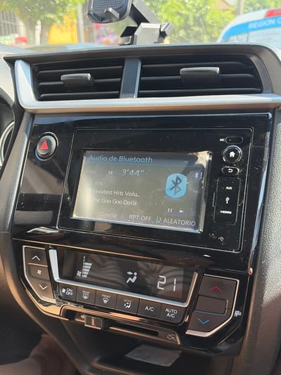 The image shows a car dashboard with an infotainment system displaying Bluetooth audio playing the song "Iris" by The Goo Goo Dolls, with a runtime of 3 minutes and 44 seconds. The system indicates that repeat is off, and random play is enabled. The climate control is set to 21 degrees, and the A/C is active. Ventilation is set to blow air to the front and the temperature is set to 21 degrees. There are physical buttons for the climate control functions and volume.
