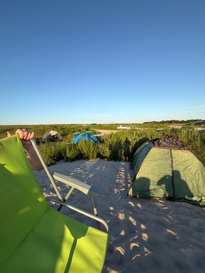 In a sandy, grassy area with a clear blue sky, a campsite is set up. Several tents of various colors, including green, blue, and beige, are pitched amongst the vegetation. Some cars are parked in the background on a dirt road. In the foreground, a person's hand is resting on the armrest of a bright green camping chair, which is positioned on the sand, facing away from the viewer. The scene appears to be a relaxed outdoor gathering.