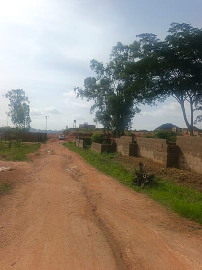 This media file depicts an outdoor scene in Reifild-Ist, Nigeria. The foreground features a dirt road with some puddles, suggesting recent rain. To the right of the road, a low wall constructed from concrete blocks is visible, with grass growing along its base. Further along the road, a white SUV is parked. In the middle ground, more block walls are under construction, and several tall trees with lush green foliage are scattered across the landscape. In the background, there are buildings, some appearing to be under construction, and a water tower. The sky is overcast, indicating a cloudy day. The overall scene suggests a developing area with ongoing construction. There is no visible text in the image.