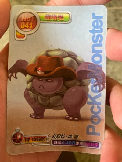 A single rectangular trading card with rounded corners, displaying surface wear including scratches and smudges, is held vertically by human hands. The left hand's fingers are visible along the left edge, and a right thumb partially obscures the card's right side. The card features a central illustration of a purple, rock-like creature resembling Golem, adorned with a reddish-brown cowboy hat that includes a small red and white Poké Ball icon. The creature has a segmented, greenish-grey shell and a scowling expression, standing on four thick, purple limbs against a background that transitions from light blue at the top to light brown at the bottom.
In the top-left corner, a red oval contains "NO: 041" in white text, adjacent to a green banner displaying Chinese characters "隆隆岩". The term "PocketMonster" is printed vertically in blue along the right edge. The bottom-left area of the card shows a circular icon and the text "HP 135920". Below the creature, Chinese text reads "必殺技:地震" (Special Move: Earthquake), followed by "身高:1.4公尺 體重:300公斤" (Height: 1.4 meters Weight: 300 kilograms). The card is presented in a well-lit environment. This scene is situated in the city of Đak Đoa, Vietnam.