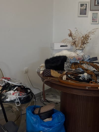 This image depicts an interior scene, likely a retail space or storage area, located in Kfar Yona, Israel. The room features plain white walls. On the upper right wall, three framed pictures are visible: one depicting a landscape with a child, another a portrait, and a third indistinct landscape. A white power outlet with a white cord plugged into it is on the lower left wall.
A dark brown, round table with a vertically fluted base occupies the right side of the frame, heavily laden with various items. Stacked white cardboard boxes, one labeled "PREMIUM," are positioned on the table against the wall. A white pitcher-style vase containing dried pampas grass and cotton bolls stands behind these boxes. On the table, a black furry hat and a white furry item are prominently displayed. The table surface is covered with an assortment of accessories, including patterned scarves (e.g., polka dot, houndstooth), multiple leather belts, and small packaged items.
On the lower left, a large clear plastic bag, filled with clothing and accessories, rests on a translucent chair or stool. On the floor beneath the table, a blue fabric tote bag (resembling an IKEA bag) contains a tan handbag and other miscellaneous objects. Sparse white material is scattered on the floor directly beneath the table. No individuals are present in the scene.