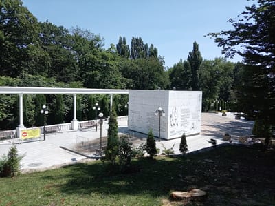 The image depicts a sunny outdoor scene in a park setting, likely in Zheleznovodsk, Russia, given the presence of Cyrillic text on the monument. In the foreground, a grassy slope with a tree stump is visible. Behind it, a paved area features several evergreen trees and a modern white cubic monument with inscribed text and images.
To the left of the monument, a white colonnade with a railing and benches runs along a paved walkway. A yellow warning sign is posted on a barrier near the colonnade. Further back, a line of tall, slender trees and dense foliage forms the backdrop. On the right side of the image, a large tree with dark green leaves partially obscures the view of a pathway and more trees in the distance. A few individuals are seated on benches in the mid-ground. The sky is clear and blue.