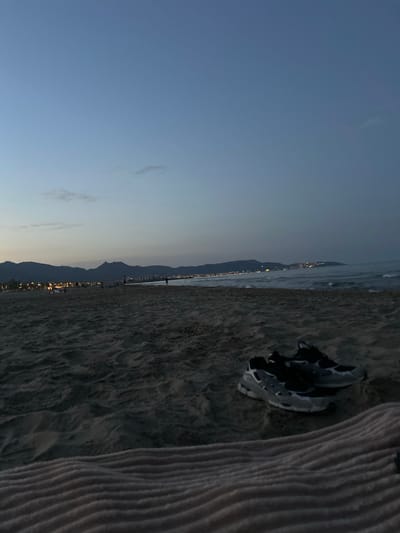 This media file shows a beach scene at dusk. The sun has set, casting a soft blue and purple hue across the sky, with a few wispy clouds visible. In the foreground, a pair of athletic shoes, predominantly grey and black with white accents, rests on the sandy beach. To the right of the shoes, a textured, striped fabric, possibly a towel or blanket, is laid out.

The mid-ground features the ocean meeting the shore, with gentle waves lapping at the sand. Beyond the ocean, a distant coastline is illuminated by a scattering of lights from a town or city nestled against a mountain range. The lights twinkle, suggesting an evening ambiance. The mountains themselves are dark silhouettes against the fading light. The overall mood is tranquil and serene, evoking a sense of peaceful closure to the day. No text is visible in the image.