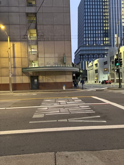 The image depicts a street scene in San Francisco, California, United States of America, likely at dusk or early evening. In the foreground, a crosswalk with white stripes and a red line is visible on asphalt pavement. In the middle ground, a building with a light-colored facade and a curved awning with "FIFTH & MISSION" signage is present. An "EXIT ONLY" door is visible on this building, as well as a blue fire hydrant. To the right of this building, a modern, multi-story skyscraper with a dark facade and numerous illuminated windows stands tall. Further to the right, another white building with traffic signals and a pedestrian crossing light showing a green figure is visible. A black car is parked on the street to the right. Several pedestrians are walking on the sidewalk near the "FIFTH & MISSION" building and in the distance. Streetlights and overhead wires are also present in the scene.
