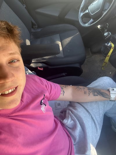 The image shows a person sitting in the driver's seat of a car. The person is wearing a pink t-shirt and grey sweatpants. They have short, brown hair and blue eyes. There are tattoos on their arm. The car's interior is visible, including the steering wheel, gear shift, and seats. The background appears to be outdoors, with sunlight casting shadows. There is a receipt on the person's lap. The image was taken in the city of Lovets, Bulgaria.