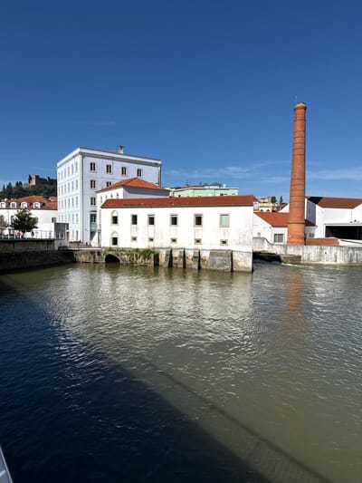 This image captures a riverside scene in Tomar, Portugal, under a clear blue sky. The River Nabão occupies the lower portion, bisected diagonally by a dark shadow, likely from a bridge, leaving the left side of the water dark and the right side lighter and reflective.

On the left bank, a multi-story building with a light blue-white facade and numerous uniformly spaced rectangular windows stands prominently. Behind it, on a tree-covered hill, the ruins of a historic castle are visible in the distant background. Adjacent to the river on this bank is a lower white structure with a red tile roof, featuring several arched openings at its base that appear to be former water channels, with some vegetation growing on the stone foundations. Faint, indistinct figures of people are visible on a walkway further left, behind a railing.

On the right bank, a long, white building with a red tile roof extends along the river, also displaying multiple rectangular windows. To its right, a tall, cylindrical industrial chimney, constructed from reddish-brown brick, rises against the sky. A small weir or waterfall is visible between structures on the right, where water flows into the main river channel. Another white building with a dark, open bay and a red tile roof is present further to the right. The scene is brightly illuminated by direct sunlight. No specific actions or interactions between individuals are clearly depicted.