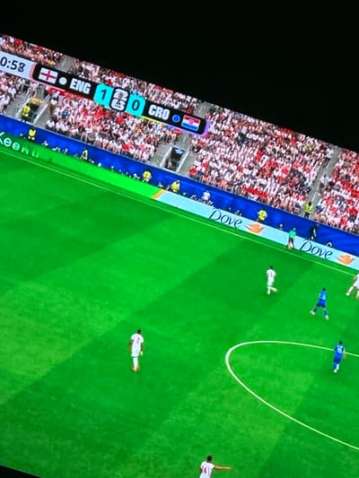 This is a high-angle, medium shot of a soccer game being played on a vibrant green field. A scoreboard at the top of the image shows "0:58" and a score of "ENG 1 0 CRO". The stands are filled with spectators wearing a mix of red and white attire. The field itself is marked with white lines. Several players in white and blue uniforms are visible on the field. Two players in white are in the foreground. One is facing away from the camera, and the other is further up the field. A player in a blue uniform is also in the foreground, facing the camera. Another player in blue is further up the field. Advertising boards with "Dove" are visible along the sideline. The scene appears to be outdoors, and the lighting suggests it is daytime. The city of Rayfield, Nigeria, is not depicted in this image.