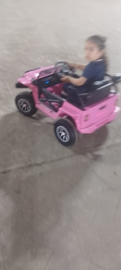 A young girl with long hair drives a pink toy Jeep on a paved surface. She wears a dark blue shirt and dark pants. The Jeep has black tires with silver rims and a black seat. Motion blur suggests the Jeep is moving. The background is a plain, light-colored pavement or concrete area. The overall image has low lighting and appears to be taken with a phone.