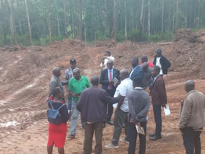 The image shows a group of approximately fifteen people standing on muddy ground in Moticho ward, Kenya. Behind them is a freshly cleared area of mud, with tree stumps and tire tracks visible. In the background, a dense forest rises. The people are dressed in a mix of casual and formal attire, including shirts, trousers, dresses, suits, and jackets. One woman wears a red skirt and carries a small backpack. One man wears a green shirt, and another a suit with a red tie. Some individuals hold papers or books. They appear to be engaged in conversation or observation. Some are interacting with one another, while others stand apart. One person in the background is holding a phone or camera.