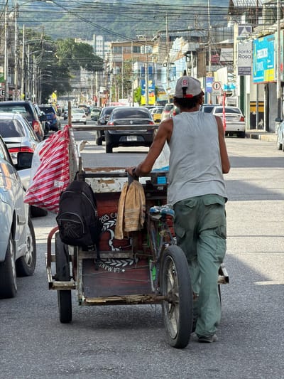 The image depicts a street scene in Maracay, Venezuela, with a man pushing a cart loaded with goods. The man, wearing a grey tank top and green cargo pants, is seen from the back as he walks alongside a line of parked and moving vehicles. His cart is laden with a red and white striped bag, a black backpack, and a beige towel. The cart itself appears to be constructed from salvaged materials, with a wooden box and a bicycle wheel.
The environment is characteristic of an urban area in Venezuela, with dense overhead power lines crisscrossing the sky. Buildings line the street, some displaying colorful signs and advertisements, suggesting a commercial district. The road is asphalt, and traffic appears to be moderate, with several cars visible. The backdrop includes a hillside with vegetation and more buildings, indicating the city's topography. The lighting suggests it is daytime, possibly late morning or afternoon, given the shadows.