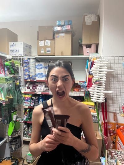 This is a vertically oriented, slightly blurry image of a woman in a store. The woman is in the foreground, looking at the camera with a surprised expression and her mouth open. She is wearing a black top and has a tattoo on her arm. She is holding a chocolate bar.

Behind the woman are shelves filled with various products. These include packages of tissues, cleaning supplies like Ariel laundry detergent, gardening tools such as trowels and rakes, and other assorted items in packaging. Some of the products have visible text in both Czech and English. The shelves are made of metal grating.

The store appears to be a retail environment, possibly a supermarket or a discount store, stocked with household goods. The lighting suggests it's daytime, but it's difficult to determine the time of day precisely. The overall impression is that the woman is posing for a photo while browsing or shopping in the store.