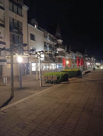 The image depicts a night scene on the Rhine promenade in Emmerich, Germany. The camera faces down a paved walkway lined with mature trees with globe-shaped canopies and built-in street lamps. To the left are apartment buildings of varying heights with windows and balconies. To the right, a hedge separates the walkway from a restaurant terrace with red parasols. A bench is visible near the hedge in the foreground. The area is illuminated by street lights and light emanating from the restaurant windows. The sky is dark, and the lighting creates shadows on the pavement. The scene is devoid of pedestrians or other visible subjects.