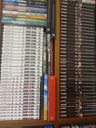 This is a media file featuring a collection of books and DVDs, likely a personal library or a display in a store. The subjects are primarily book spines and DVD cases, with the main objects being manga volumes from series like "Attack on Titan" (L'ATTACCO DEI GIGANTI), "Vinland Saga", and "Tokyo Ghoul". There are also DVD cases for "Fate/stay night [Heaven's Feel]" and "Just Dance 2020".

The scene is set on wooden shelves, suggesting an indoor environment like a home or a shop. There are no people visibly depicted. The activity taking place is simply the presentation of these media items, with no explicit event or action occurring within the frame.

Notable details include the titles and numbering of the manga volumes, indicating a comprehensive collection of certain series. The "Attack on Titan" volumes are numbered from 1 to 34, and "Tokyo Ghoul" volumes are numbered from 1 to 16. The "Vinland Saga" volumes are also numbered, from 1 to 8. The "Fate/stay night" DVDs are also present. The spine of one DVD case appears to be labeled "unlimited blade works." The overall impression is one of organized collection, with a focus on Japanese manga and related media. The presence of Italian text on some spines, such as "L'ATTACCO DEI GIGANTI" (Attack on Titan) and "Star Comics," suggests the location context of Monsummano Terme, Italy. The lighting appears to be artificial, typical of an indoor setting, and there are no indications of specific time of day or weather conditions. The emotions conveyed are neutral, due to the absence of human subjects.
