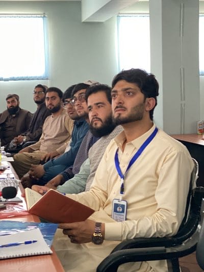 A group of men sit in a room, presumably at a table. They appear to be attentive, and the man closest to the camera holds a book. One man has a lanyard with a name tag around his neck. A notebook and pen sit on the table. The location is assumed to be Shurandam, Afghanistan.