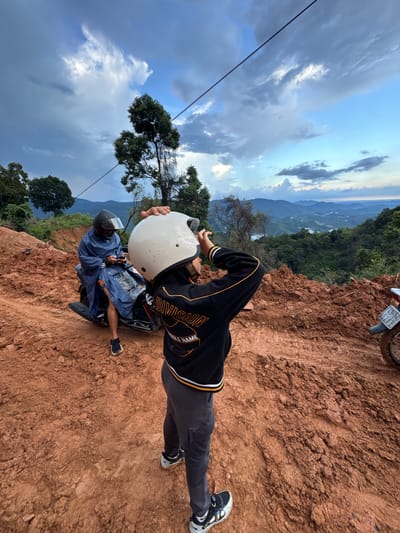 This is a medium shot, full-color, outdoor photograph taken from a low angle, looking up at two people and a scenic mountain landscape. The foreground is dominated by a muddy, reddish-brown dirt road, with tire tracks visible. To the left, a person wearing a blue rain poncho and a black helmet sits on a dark scooter. They are looking down at their phone. In the center and right foreground, another person wearing a black and yellow jacket and a white helmet stands with their back to the camera, holding the helmet up to their head as if to take a photo. The background features a lush, green mountain range with several layers of hills receding into the distance, under a sky with dramatic clouds and hints of blue. Power lines are visible stretching across the sky. The scene appears to be on a mountain road, possibly in a rural area. While the image does not explicitly depict Nao Sẻ, Vietnam, the landscape is consistent with mountainous regions in Southeast Asia.