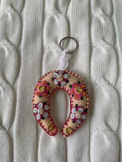 A handmade, horseshoe-shaped keychain lies on a white, cable-knit surface. The keychain is primarily beige felt, decorated with pink embroidered flowers, shiny silver sequins, and small gold beads. A pink embroidered trim borders the edge. The keychain is attached to a silver metal key ring by a short length of white, ribbon-like material. The background is a heavily textured, white cable-knit fabric. The scene is likely indoors in Лесной Пейзаж Кп (шарапово, Russia, based on the image name.