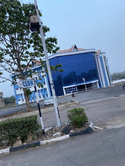This image depicts an outdoor daytime scene on what appears to be a university campus in Osu, Nigeria, under an overcast sky. The foreground shows an asphalt road with black and white painted concrete curbs, bordering sparse green bushes and exposed earth.
A tall, grey metal pole stands prominently in the left-center, featuring a rusty, square electrical box mounted at its upper section. Attached lower on this pole is a white directional sign with a blue arrow pointing left, inscribed with "CEDM" in blue capital letters. A smaller, black and white striped pole is positioned next to it. A tree with green foliage is situated behind the pole, partially obscuring a building to the left.
In the midground, to the right, is a multi-story building characterized by expansive dark blue reflective glass facades, framed by white and light blue structural components, and topped with a brown gabled roof. A sign above its main entrance reads "FACULTY OF LAW" in white capital letters, with a light-colored statue or emblem directly above it. Two individuals are present near this building's entrance: one person in a red shirt and dark trousers walks towards the building's steps, while another person in dark clothing walks away from the entrance towards the right side of the frame.
To the left of the "FACULTY OF LAW" building, another multi-story structure is visible, predominantly white with light blue horizontal banding and window frames, sharing a similar brown gabled roof. A silver sedan is parked on the asphalt road between these two buildings. A dark-colored vehicle is partially visible on the far right edge of the frame. Distant hills or structures are discernible on the horizon.