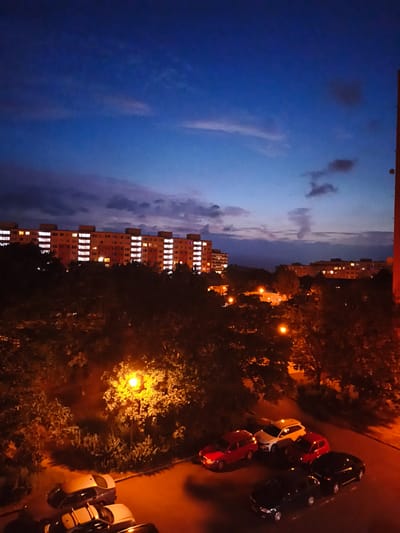 This is a medium-shot, eye-level, nighttime photograph taken from a high vantage point, likely a residential building, overlooking a street and park area in the city of Most, Czechia. The sky is a deep twilight blue with scattered clouds. In the background, several long, multi-story apartment buildings are illuminated with many windows lit from within, suggesting it is evening or night. In the mid-ground, dense trees, some with illuminated foliage from streetlights, form a dark canopy over the scene. A bright yellow streetlight illuminates a section of the trees and the pavement below. In the foreground, several cars are parked on the asphalt. These include a red sedan, a white SUV, two red hatchbacks, and two black sedans. The overall atmosphere is one of urban residential life at dusk.