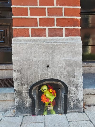 A small, green, anthropomorphic figure, possibly a toy or figurine, is positioned in an arched recess of a stone wall in front of a red brick facade. The figure is holding a bouquet of colorful artificial flowers, predominantly orange and yellow, with some red accents. The figure's head is tilted, and it appears to be smiling with its eyes closed. The recess is framed by a dark, arched border. Below the figure, there is a label with text that is partially obscured, but "TILBURG" is visible. The surrounding environment consists of a cobblestone pavement, stone steps, and a grate. The overall scene suggests an urban outdoor setting, likely a street or building entrance. The text "TILBURG" on the label indicates the location as the city of Tilburg in the Netherlands.