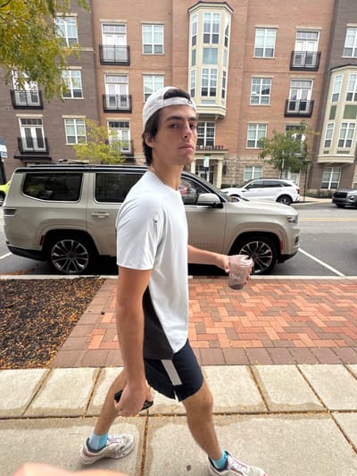 A young adult male is depicted walking on a concrete sidewalk in front of "The Foundry" apartment complex, located at 211 E South St, South Bend, Indiana, United States. He wears a light grey short-sleeved athletic shirt, dark athletic shorts with a light stripe, white athletic shoes, light blue socks, and a light-colored baseball cap worn backward. The subject holds a translucent plastic cup containing a brown liquid with a dome lid in his right hand and a black smartphone in his left hand, partially visible. His head is turned towards the viewer.
Behind the subject, a beige/tan Jeep Wagoneer is parked parallel to the street. Further down the road, a white SUV and a dark vehicle are also parked. The background features the multi-story brick facade of "The Foundry," characterized by numerous windows, some with black iron balconies, and a prominent bay window structure. Adjacent to the concrete sidewalk is a strip of reddish-brown brick pavers arranged in a herringbone pattern, bordering the street. Between the sidewalk and the building, a narrow bed contains dark mulch and scattered autumn leaves. Trees with green and yellow foliage are visible, suggesting an autumn season under diffuse daylight.