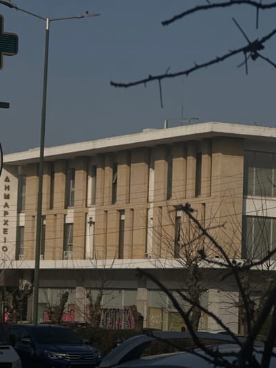 The image depicts the Agia Paraskevi City Hall in Agia Paraskevi, Greece, on a clear, bright day. The building occupies the background, presenting a multi-story facade of light tan or beige material with prominent vertical pilasters separating recessed window sections. The letters "ΔΗΜΑΡΧΕΙΟ" (City Hall) are vertically visible on the upper-left side of the building. The structure features a flat roof with a television antenna and a separate, slender antenna visible against the blue sky. Below the main three floors of the building, a ground level with large, dark-tinted or boarded windows is visible, with graffiti tags in pink, black, and white on a low wall or structure in front of it. Several air conditioning units are mounted on the building's exterior. In the midground, bare tree branches with visible thorns are heavily silhouetted in the foreground, partially obscuring the lower half of the building and the street. Utility lines traverse horizontally across the mid-section of the image. A dark green streetlight pole stands vertically on the left, next to a partial view of a green cross sign. In the lower midground, a dark blue SUV is parked or moving, with other vehicle parts visible to its left and right. Sparse green bushes are visible between the street and the building's ground level. No identifiable individuals or specific actions are clearly discernible within the frame.