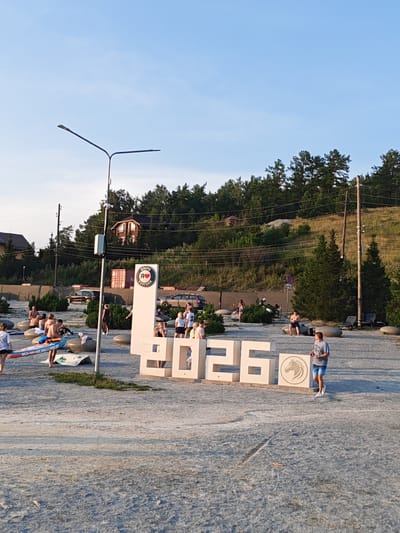 The image depicts a scene at a lakeside resort in Turgoyak, Russia, on a clear day with a bright blue sky. In the foreground, a large, white, three-dimensional sign reads "2026" with a circular emblem featuring a horse's head and the text "Я ЛЮБЛЮ ТУРГОЯК" (I Love Turgoyak) to the left of the number six. The sign is situated on a sandy or gravelly ground surface.

Scattered around the sign are various people, most of whom appear to be vacationers. Several individuals are standing, sitting, or walking. To the left of the sign, a person is holding a paddleboard. Other people are seen on or around large, smooth, grey rocks. In the background, there is a dense hillside covered with trees and bushes. Several houses are visible nestled among the trees on the slope. Power lines and utility poles are also present.

Further in the background, a few vehicles, including cars and a motorcycle, are parked. The overall environment suggests a recreational area, likely a beach or park setting near a body of water, given the presence of the paddleboard and the resort-like atmosphere. The lighting indicates it is likely late afternoon or early evening.