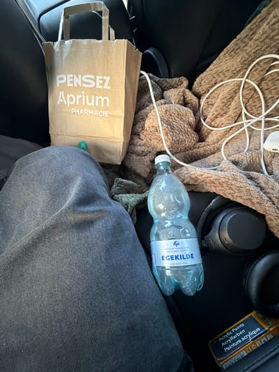 The image displays a close-up view of items likely inside a vehicle, possibly a car, given the presence of a dark, textured car seat. In the foreground, a person's dark blue denim-clad legs are visible, suggesting they are seated. To the right of the legs, a brown paper bag with white text that reads "PENSEZ Aprium PHARMACIE" is positioned.

Further to the right, a brown textured blanket is draped, with a white cable coiled on top. A clear plastic bottle of "EGEKILDE" mineral water sits on the blanket. Next to the water bottle are a pair of black Sony headphones. In the lower right corner, a small, orange and black box labeled "Acrylic Paints" is visible.

The scene appears to be indoors or within a sheltered space, with the lighting suggesting it is daytime. There is no direct indication of the specific location within the city of Ödsmål, Sweden, beyond the general context of items commonly found in a vehicle.