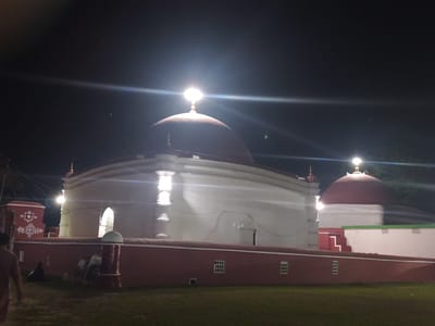The image shows a nighttime view of the Bibi Begni's Mosque in Bagerhat, Bangladesh. The mosque, an ancient structure, is illuminated by artificial lights, highlighting its white walls and the large, dark red domes. A low red wall with decorative white motifs surrounds the mosque complex. A person is seen walking away from the camera on the left side of the frame, and another person is seated near the red wall. Two bicycles are parked near a pillar in front of the mosque. The ground in front of the mosque appears to be a grassy area. The sky above is dark, suggesting it is nighttime.