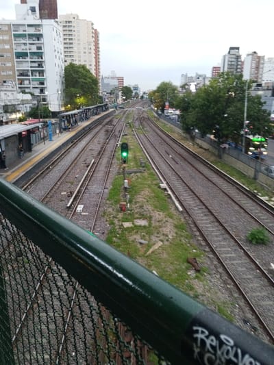 The image depicts an elevated view of the Ramos Mejía train station and adjacent urban landscape in Ramos Mejía, Argentina, under an overcast sky or dim lighting conditions.
In the foreground, a dark green metal handrail with "Revol" graffiti and a green chain-link fence partially obscures the lower portion of the scene, indicating the vantage point is from an overpass or bridge.
The midground features multiple parallel train tracks, approximately five to six visible, converging into the distance. Between tracks, gravel, soil, and patches of green vegetation are present. A prominent signal pole with an illuminated green light is positioned between two central tracks. To the left, a train station platform with a yellow safety strip and multiple red-roofed shelters is visible. Several individuals, estimated between five and ten, are standing on the platform, waiting. To the right of the tracks, a multi-lane road runs parallel, populated with moving vehicles, including a green and white bus with illuminated headlights. Trees line this road, partially obscuring the view.
The background consists of a dense urban environment. On the left, tall, light-colored multi-story residential buildings with numerous balconies dominate the skyline. Further right, more diverse urban structures, ranging in height, extend towards the horizon under a uniformly light gray sky.