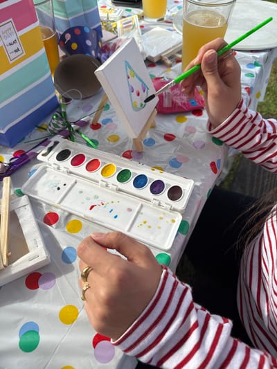 An overhead view captures an outdoor art activity taking place on a table in Accord, United States. The scene is illuminated by bright sunlight. In the foreground, a person, wearing a red and white striped long-sleeved shirt and gold rings on their left hand, is actively painting a small white canvas mounted on a miniature wooden easel. The right hand holds a green-handled paintbrush, applying green paint to an outlined party hat design on the canvas, which already features multicolored polka dots. An open white plastic watercolor paint set, containing multiple colored paint pans (black, red, yellow, green, blue, purple, brown) and mixing wells with paint splatters, rests on the table. The table is covered with a clear plastic tablecloth adorned with a scattered multicolored polka-dot pattern. Nearby objects include a blue and white striped gift bag labeled "happy day! TO NOAH FROM RGR," two glasses containing an amber liquid beverage, a brown spherical object, and green tinsel. Another small, unpainted canvas on a miniature easel is visible to the left. Green grass is discernible in the soft-focus background.