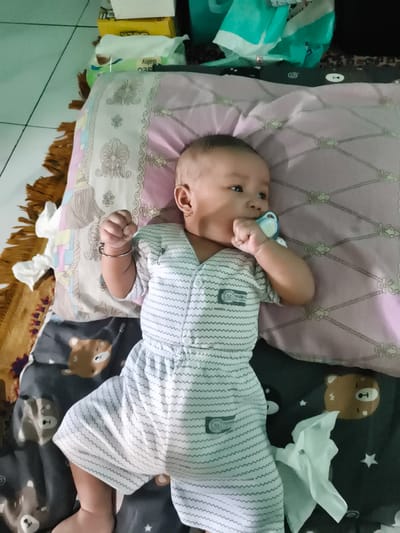 A baby lies on a patterned blanket with cartoon bears and stars. The baby is wearing a white onesie with gray wavy stripes and a light gray pacifier in their mouth. On their left arm, they wear a thin silver bracelet. The baby is looking to the right with a neutral expression. Behind the baby, a pink pillow with a rope and floral pattern is visible, along with stacked boxes and packaging. The setting appears to be indoors, with a tiled floor visible to the left and what looks like a crib or bed. The time of day and weather are not discernible.