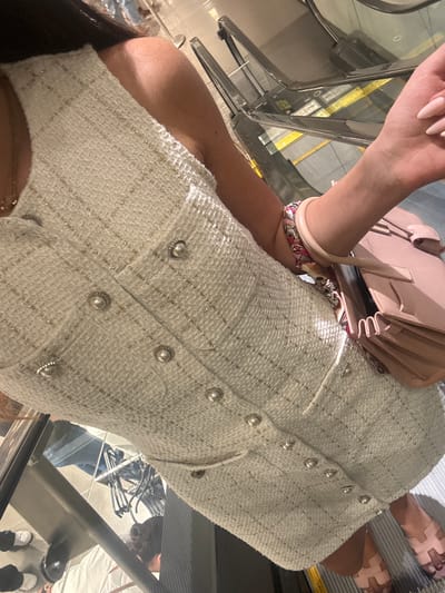 A person stands on an escalator in a brightly lit indoor environment. They are wearing a sleeveless white tweed dress adorned with pearl buttons and gold threads. A pale pink handbag is held in their right hand, and a colorful patterned bracelet is worn on their left wrist. The person's left arm is visible from the shoulder down to the wrist. The background shows the metallic structure and steps of the escalator. The location is Aventura, United States of America.