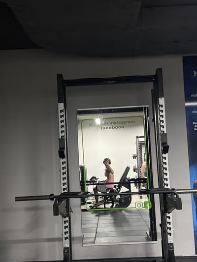 This image captures a reflection in a gym mirror, showing a person engaged in a workout. The primary subject is a person, likely a woman given the pink athletic shorts and athletic top, performing an exercise. The setting is a gym, evidenced by the presence of exercise equipment, including what appears to be a weight rack or a similar apparatus with multiple pegs for holding weights or attachments. The reflection also shows a wall with vertical wooden panels, characteristic of some gym environments.

A green graphic overlay is visible within the reflection, featuring social media icons for Instagram and Facebook, along with text that reads "on Instagram" and "facebook". This suggests the gym may promote its social media presence. The lighting is typical for an indoor setting, suggesting it's likely daytime or evening within the gym's operational hours. There are no clear indicators of time of day, weather, or specific emotions expressed by the individual. The image is oriented vertically, implying the photographer was holding their phone in portrait mode. The overall scene is utilitarian and focused on fitness activities.