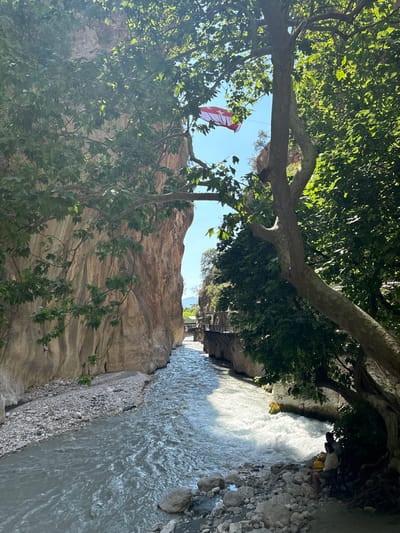 The image shows a view from within a canyon, looking down a flowing river. On the left side of the canyon, a sheer rock face rises vertically. On the right side, there is a wooden walkway built along the canyon wall, with trees and foliage overhead. In the distance, a bridge spans the canyon. In the foreground, two people are sitting on rocks near the riverbank, under the shade of a large tree. A Turkish flag is visible flying from a wire strung across the canyon, high above the river. The river itself appears to be fast-flowing with some whitewater. The scene is likely the Saklıkent Gorge, located near the town of Kaş, Turkey.