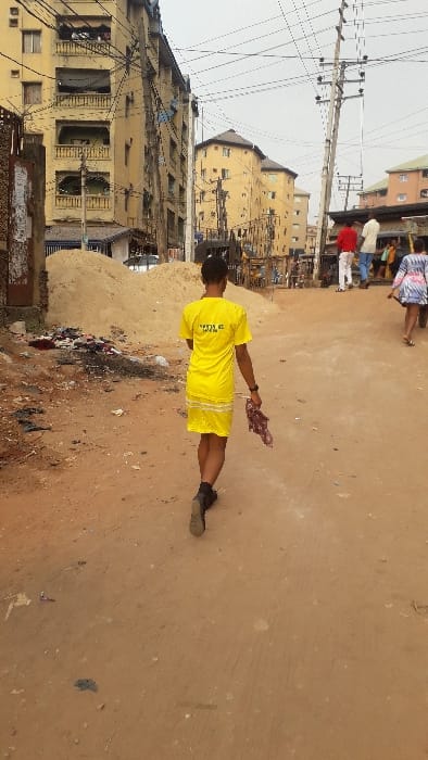 A person in a yellow outfit stands in the middle of a dirt road in a somewhat urban area. They hold a piece of cloth in their right hand. Buildings line the street, and power lines crisscross overhead. A pile of trash sits on the side of the road. Other people are visible in the distance at the top of the image. The road is light brown and dusty, and the buildings appear old and weathered.