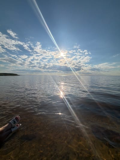 The image shows a person's feet wearing white slides with a green crocodile logo, submerged in shallow, clear water. The water is illuminated by sunlight, creating rippling patterns and reflections on the sandy bottom. The perspective is from the water level, looking upwards towards the sky and horizon.

The scene is a body of water, likely a river or lake, with a shoreline visible in the distance. The sky is bright blue with scattered white, fluffy clouds. The sun is shining brightly, casting lens flares across the image. The time of day appears to be late morning or early afternoon, given the intensity of the sunlight and the angle of the sun. The weather is clear and sunny.

The overall mood is peaceful and relaxing, suggesting a leisurely activity like wading in the water on a warm day. The location context provided is Vyshhorod, Ukraine, which is known for its Dnieper River and reservoir. The image captures a serene moment of enjoying nature by the water.