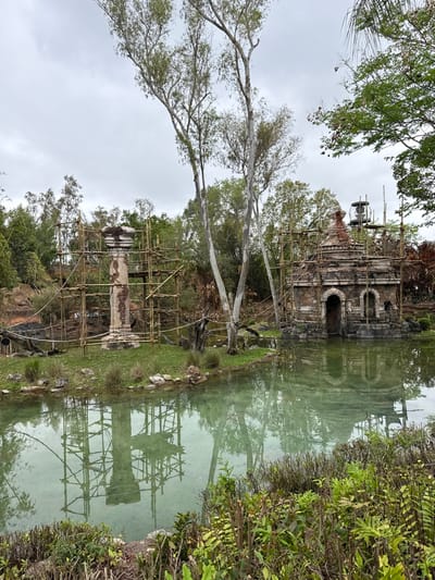 This image depicts a tranquil scene in Bay Lake, Florida, United States, specifically within Disney's Animal Kingdom theme park. Under an overcast sky, a placid body of water occupies the foreground and midground, reflecting the surrounding environment.

Two distinct structures, both enveloped in extensive bamboo scaffolding, are situated on small land masses within or bordering the water. To the left, a tall, light-colored column or pillar, exhibiting ornate carving on its capital and base, stands upright. Its shaft shows signs of weathering or discoloration. Bamboo scaffolding completely encases this column, with horizontal and diagonal poles visible. Rope or cable barriers are strung near the water's edge on this island.

To the right, a stone-faced building designed to resemble an ancient temple or ruin rises from the water. Its facade features an arched wooden doorway and two arched, empty niches. The building has a multi-tiered roof culminating in a conical structure. Similar to the column, this temple-like building is also surrounded by a dense bamboo scaffolding network. Behind and slightly above the temple, partially obscured by trees and scaffolding, a metal utility structure with a circular top is visible.

The landscape includes lush green foliage in the foreground, with dense trees and vegetation in the background. Several large, tall trees with light-colored bark and sparse foliage are prominently positioned in the center of the image. The calm, pale green water clearly mirrors the structures and trees. No individuals are present in the scene. The scaffolding indicates ongoing construction, refurbishment, or maintenance work on both structures.