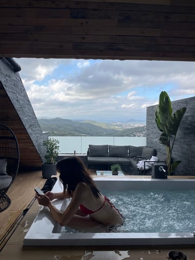 A woman in a red bikini sits in a hot tub on a wooden deck with a view of a hilly landscape. She is holding a smartphone in her hands. The hot tub is filled with bubbling water. In the background, there is an outdoor lounge area with a gray sectional sofa and a large potted plant. The scene is set on a balcony or patio of a building with a sloped wooden ceiling and a stone-clad wall. The landscape in the distance appears to be a town nestled amongst green hills, suggesting the location is Can Güell, Spain. The sky is partly cloudy.