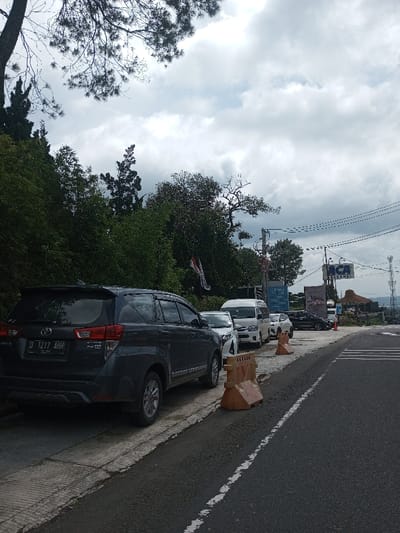 The image depicts a roadside scene in Gubbisamamidi, India, under an overcast sky. A dark grey Toyota Innova SUV, bearing license plate D 1217 AHH, is parked on the far left of the frame, partially on a concrete sidewalk, facing away from the viewer. Behind this vehicle, a white sedan and a white minivan are also parked in a continuous line on the sidewalk. Several orange plastic traffic barriers are strategically placed along the edge of the parking area, delineating it from the asphalt road. The road exhibits a solid white edge line and a partial dashed white lane marker. To the immediate left, a steep green incline densely covered with varied foliage and trees ascends. In the middle and background, multiple utility poles support an array of overhead wires. Prominent commercial signage includes a blue and white "ACA ASURANSI" sign and another darker billboard with white text, partially visible, suggesting "EXPRESSO". A building featuring a distinct peaked roof is discernible in the distance near the "ACA ASURANSI" sign. No individuals are visible within the scene.
