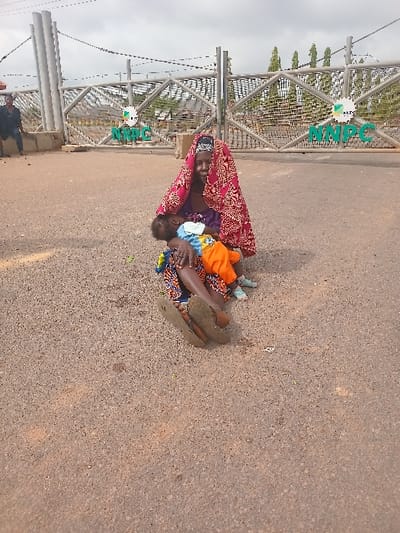 The image shows a woman and a young child sitting on the ground in front of a metal gate. The woman is wearing a red and white headscarf and is holding the child in her arms. The child is wearing an orange shirt and blue pants. The gate is closed, and there is a sign on it that reads "NNPC". The scene is in front of the NNPC Gate in Ungwan Tanko, Nigeria, located at 10.422° N, 7.49° E. The environment is dusty and dry, and the ground is made of gravel. The woman and child appear to be waiting or seeking assistance. There is a man visible in the background, but there is no interaction between him and the woman and child.