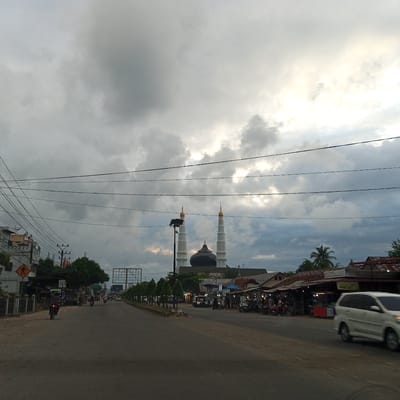 This image captures a street scene in Pantegajah, Indonesia, under an overcast sky with patches of brighter light visible through the clouds, indicating either sunrise, sunset, or partial clearing. The central background features a prominent mosque with a large, dark, possibly black or dark green dome, flanked by two tall, white minarets topped with gold-colored conical spires.

In the middle ground, an asphalt road stretches into the distance. Several vehicles are present: a white multi-purpose vehicle (MPV) or minivan is observed on the right side of the road, partially blurred due to motion, suggesting it is moving towards the viewer. Further down the road, multiple motorcycles are visible, with at least one rider in the foreground on the left, moving away. On the left side of the road, there are buildings, power poles, and a dense network of overhead electrical and communication wires spanning across the upper half of the image. A yellow pedestrian crossing sign is visible on the left.

The right side of the road features a row of small, tree-like plants lining the median, followed by various roadside structures that appear to be small shops or stalls, some with corrugated metal roofs and illuminated interiors, suggesting commercial activity. People are gathered around these stalls, some on motorcycles, indicating typical street market or daily commuter activity. Palm trees and other vegetation are scattered throughout the background and along the sides of the road. A street light fixture is visible near the mosque. The overall lighting suggests either early morning or late afternoon, given the atmospheric conditions.