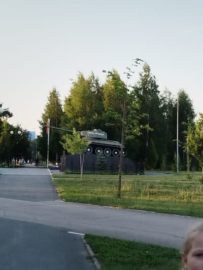 The image shows a memorial with a Soviet-era tank on a pedestal. The text on the tank reads "МАТЬ - РОДИНА" (Motherland). The memorial is located in a park or public space with trees and grass. A paved walkway is visible in the foreground, and a few people are walking in the background. The sky is clear and light. The scene appears to be set in Roshchinsky, Russia, as suggested by the text on the tank.