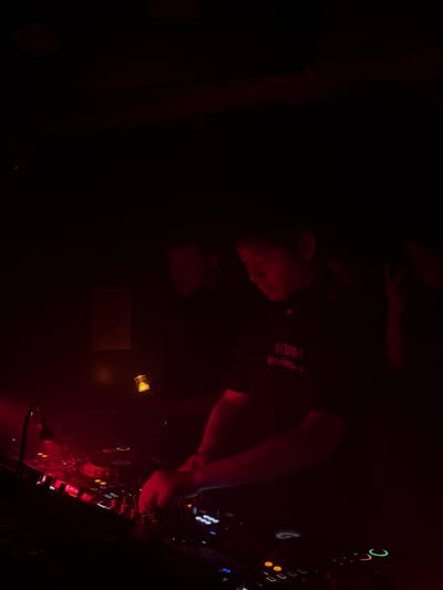 The image depicts a dimly lit nighttime scene within an entertainment venue located in Changning District, China. The primary subject is a male DJ positioned in the midground, illuminated by strong red stage lighting. He is wearing a dark t-shirt with light-colored text partially visible on the chest, reading "NCUM 1st anniversary." Headphones are resting around his neck. Both of his hands are engaged with a professional DJ mixing console in the foreground, manipulating various faders, knobs, and buttons that emit red, green, and orange light. To his left, a black gooseneck microphone is positioned near the equipment. Another individual is partially visible in the blurred background to the DJ's left, standing behind him. A third, highly indistinct figure or arm is barely discernible on the far right edge of the frame. Above the equipment, slightly to the left, a small, yellowish-glowing object, possibly a bottle containing a light source, is visible. The background consists of dark, indistinct areas, with subtle hints of architectural features or fixtures. The overall environment is characterized by dark shadows and intense red illumination.