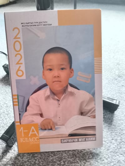 This is a photograph of a school-related item, likely a certificate or report card, featuring a young boy. The boy is the central subject, positioned in the middle of the frame. He appears to be around 6-8 years old, with short dark hair and a fair complexion. He is wearing a light pink collared shirt. His expression is neutral, and he is looking directly at the camera. His right hand is resting on an open book, with his index finger pointing towards it, suggesting he is either studying or showing something in the book.
The item itself is vertically oriented and appears to be a printed document with a photograph inset. The background of the photograph is a light blue with some stylized elements. The text at the top of the document, in a Cyrillic script, reads "№52 КЫРГЫЗ-ТҮРК ДОСТУГУ ЖАЛПЫ БИЛИМ БЕРҮҮ МЕКТЕБИ," which translates to "№52 Kyrgyz-Turkish Friendship General Education School." To the left of the photograph, a large vertical "2026" is printed in orange.
At the bottom of the document, there is another section with text. A prominent orange square contains the text "1-A КЛАСС" in white, indicating the class or grade level. Below this, in a gray rectangular box, is text in Cyrillic that reads "БИРИНЧИ МУГАЛИМ," which means "First Teacher."
The overall setting appears to be indoors, as suggested by the lighting and the background, which seems to be a carpeted floor. The image is taken from a slightly elevated angle, looking down at the document. The lighting suggests it is daytime. There are no specific cues for weather or time of day beyond the ambient light. The document seems to be placed on a textured surface.