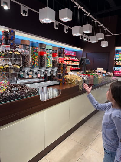 The image shows a young girl in a blue sweater and jeans standing in front of a candy store, likely in Sokilnyky, Ukraine, as evidenced by similar stores in the region. She is looking at a wide selection of colorful candies and chocolates displayed in transparent bins and on shelves behind a counter. The store appears well-lit with modern hanging lights. The girl's right hand is raised and extended towards the candy display, suggesting she is reaching for or pointing at something. In the foreground, several clear plastic cups are stacked, ready for customers to fill with their chosen treats. The store's interior features dark wood accents on the counter and light-colored cabinetry below. The floor is tiled in a neutral tone. The overall scene depicts a typical retail environment for confectionery.