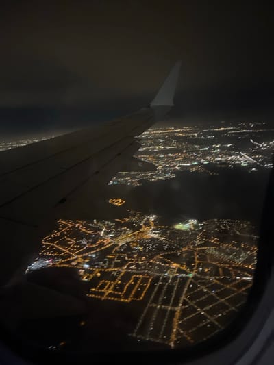 A night aerial view from an airplane window shows the wing and cityscape lights. The city lights are blurred, forming grid-like patterns and illuminated lines. Part of the plane's wing is visible in the lower left corner, casting shadows. The window frame is partially seen. Outside the window, the dark sky contrasts sharply with the bright city lights.