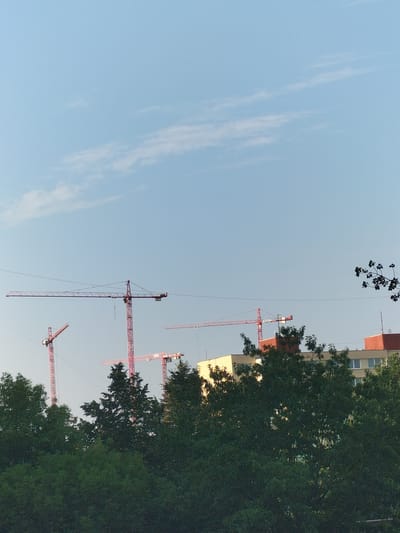 The image captures a daytime scene in České Budějovice, Czechia, featuring a clear sky with sparse clouds. Dominating the upper portion of the frame are several large construction cranes, their red structures contrasting with the pale blue sky. In the middle ground, the upper floors of multi-story buildings are visible, painted in light yellow and red. Dense green trees fill the foreground and parts of the middle ground, obscuring the lower levels of the buildings and the ground. The overall impression is that of an ongoing construction project in an urban environment, partially overgrown with vegetation. There are no people visible in the image.
